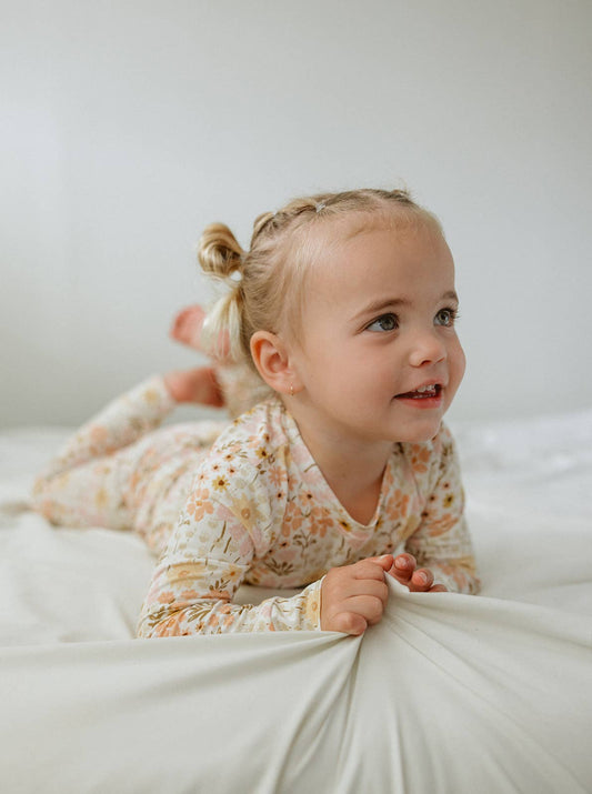 A bright, lifestyle photograph of a young girl with blonde hair styled in two small buns on her head. She is lying on her stomach on a clean, off-white sheet, looking off-camera with a gentle, curious expression. She wears a long-sleeved bamboo two-piece pajama set featuring a vintage-inspired floral print with peach, muted orange, and mustard yellow wildflowers. Her hands are lightly gripping the soft, draped fabric off the sheet in the foreground.