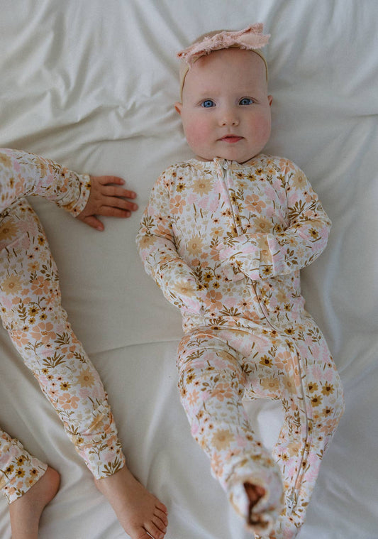 A soft, lifestyle photograph of a baby lying on a clean, white textured sheet. The baby is wearing a long-sleeved bamboo romper featuring a warm, vintage floral print with shades of peach, apricot, and muted yellow wildflowers. A delicate, dusty pink bow headband is worn on the baby’s head. The image is shot from a top-down angle in soft, natural light, highlighting the gentle, comfortable nature of the fabric.