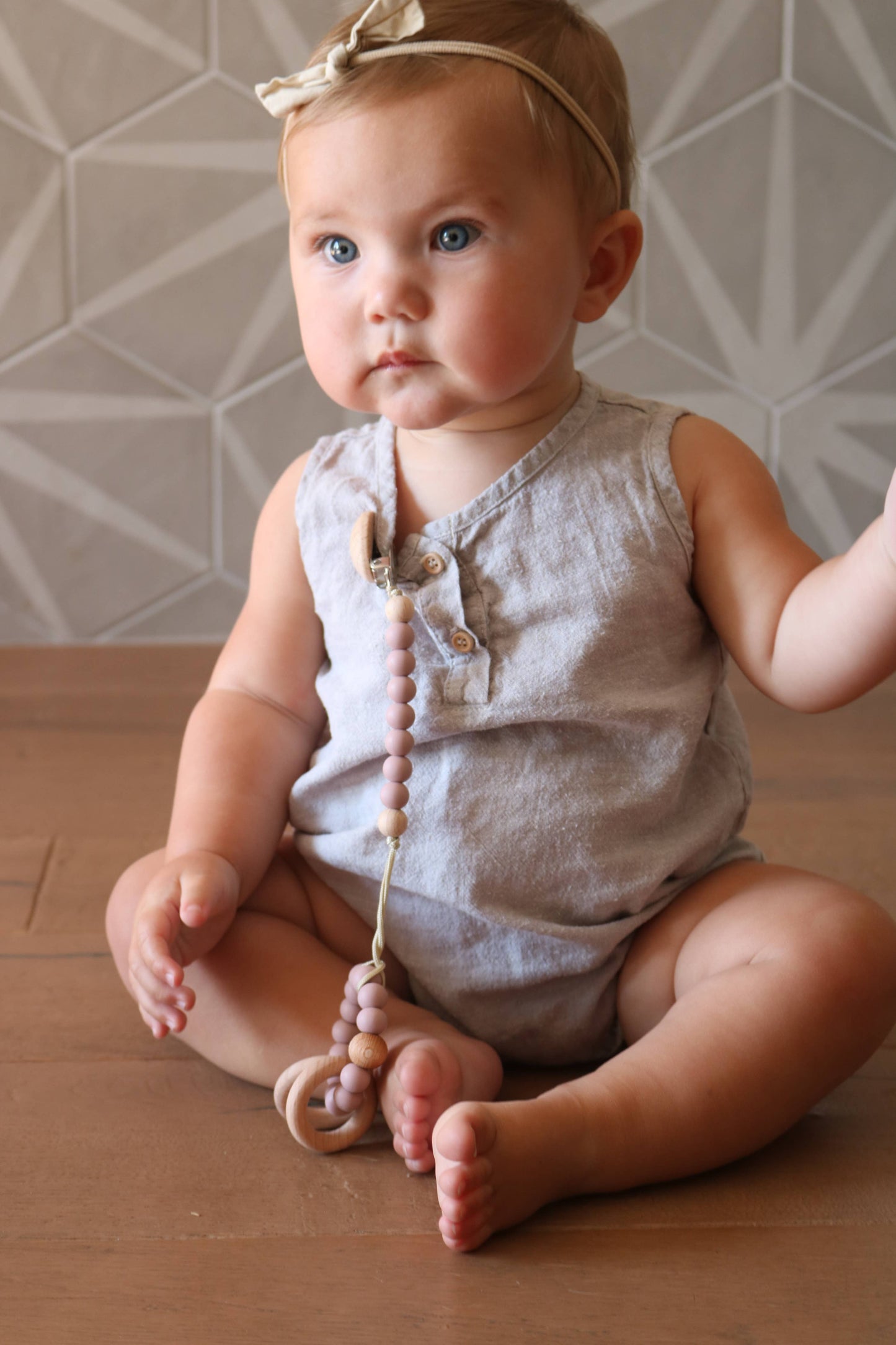 Toddler wearing a grey linen romper and bow headband, sitting on a wood floor with a silicone and beech wood pacifier clip attached to their outfit.