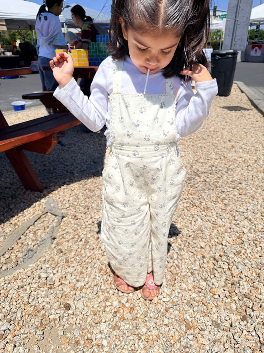 Child wearing a patterned overalls standing on a gravel surface with people and tents in the background.
