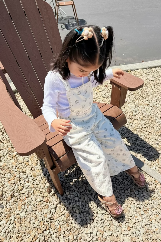 Child in a white dress with floral patterns standing on a brown chair outdoors.