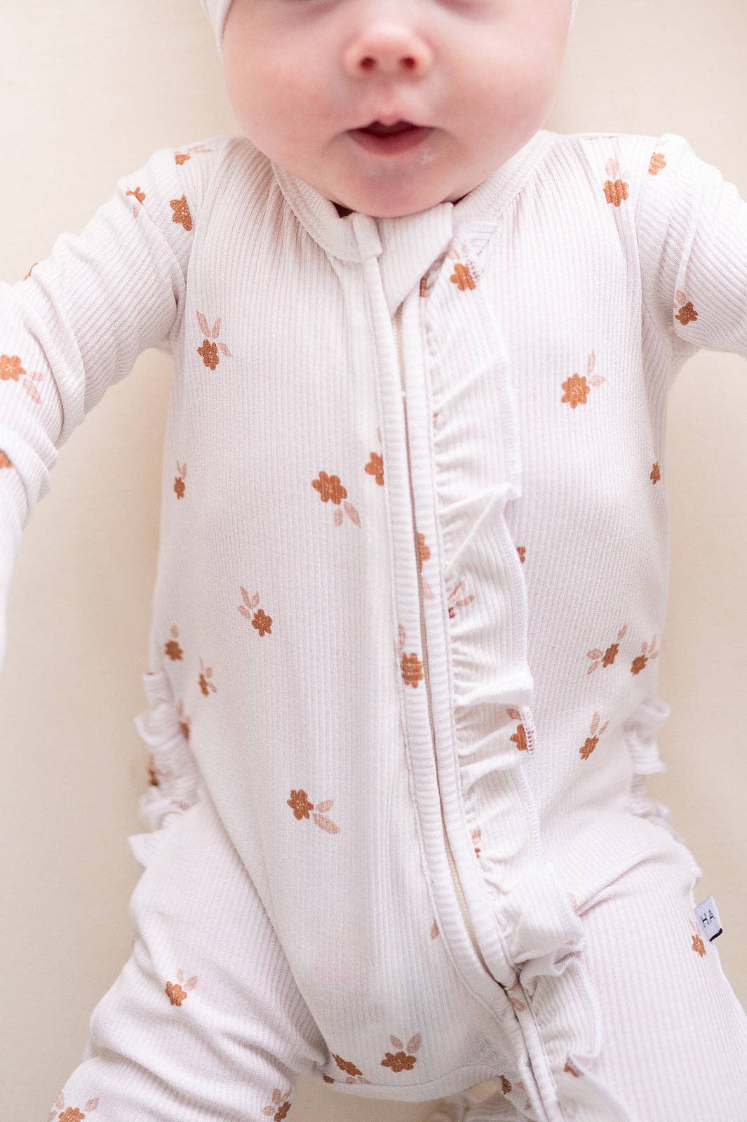 A close-up of a baby wearing a white ribbed long-sleeve footless sleeper with a front zipper and small brown floral print, set against a plain light beige background.