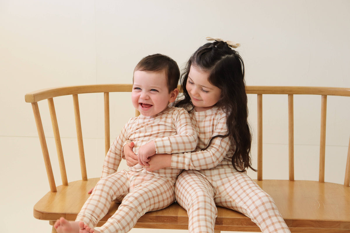 Two children sitting on a wooden bench wearing matching beige and white checkered long-sleeve pajamas.