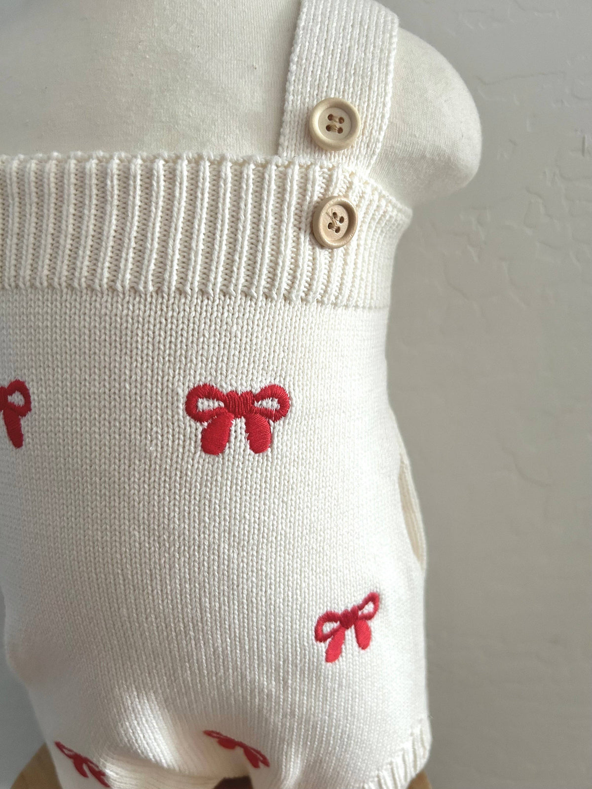 A close-up of a cream-colored knit romper or bodysuit displayed on a mannequin torso. The garment features red embroidered bow motifs and two light wood buttons on the shoulder straps.