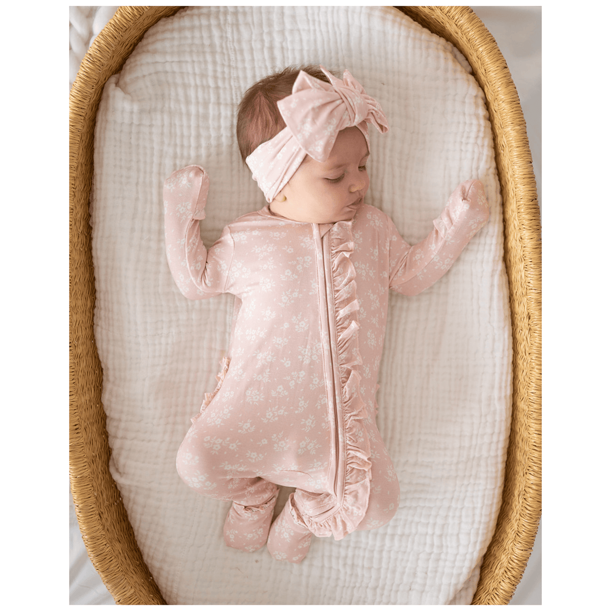 Baby in a pink outfit and headband lying in a crib with white bedding.