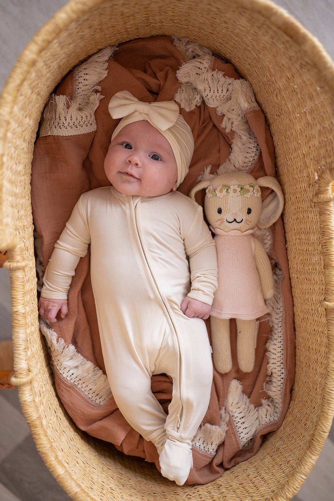 A lifestyle photograph of a baby lying in a woven wicker Moses basket on a terracotta colored fringed blanket. The baby wears a solid cream-white bamboo zipper romper and a matching knotted bow headband. Next to the baby is a knit bunny plush toy wearing a floral crown. The scene is bright and warm, emphasizing the soft, premium feel of the bamboo fabric.