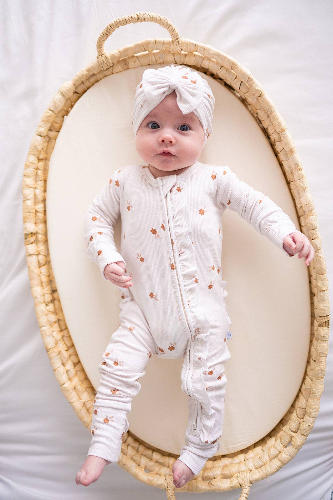 A baby wearing a white long-sleeve footed onesie with small orange floral prints and a matching white headband, posed inside a woven oval basket on a white fabric background.