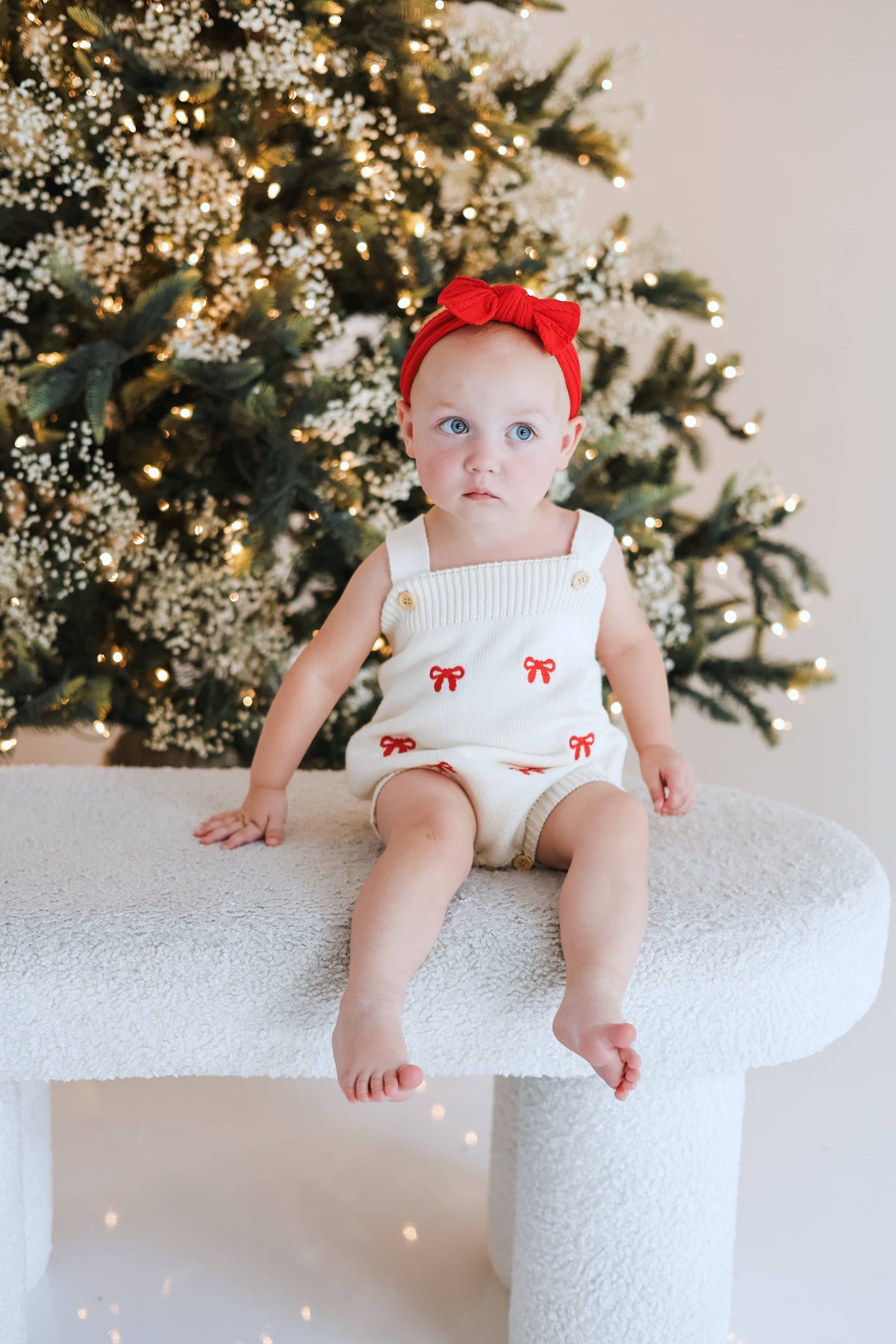 A baby girl wearing a white ribbed knit holiday romper with red bow details and a matching red bow headband, sitting on a white cat tree in front of a decorated Christmas tree.