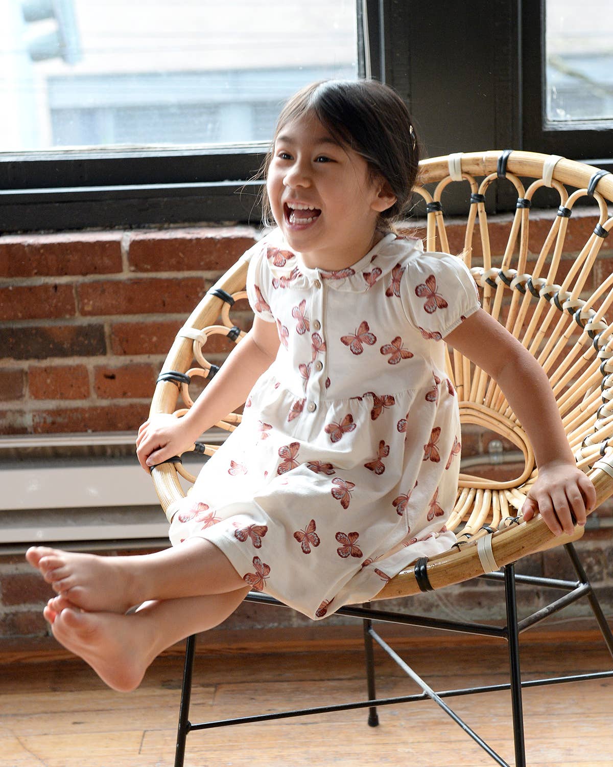 Child in a white dress with pink butterfly pattern sitting on a wicker chair indoors.