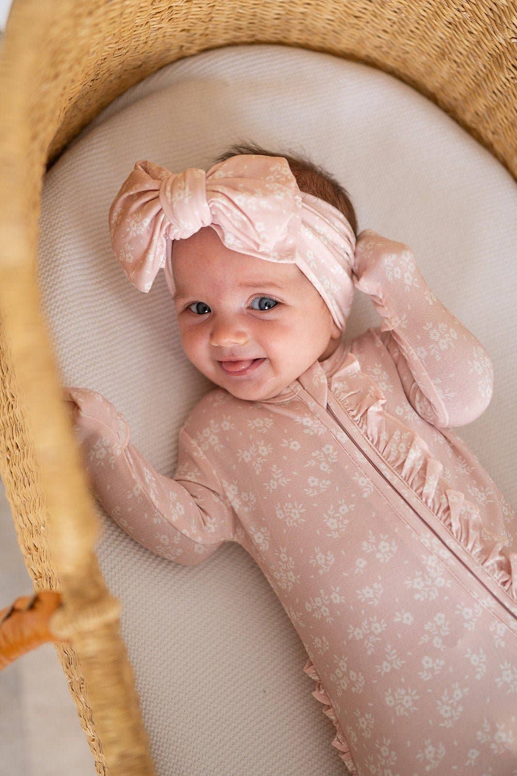 Baby in a pink floral outfit with a bow headband lying in a wicker basket.