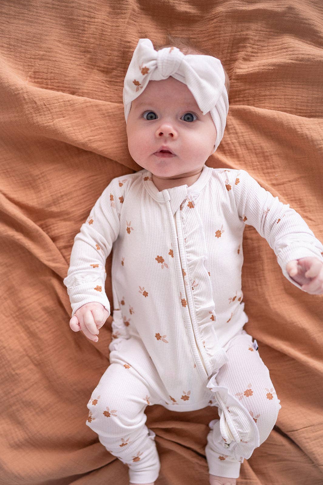 Baby in a white onesie with small patterns, sitting on a brown textured blanket.