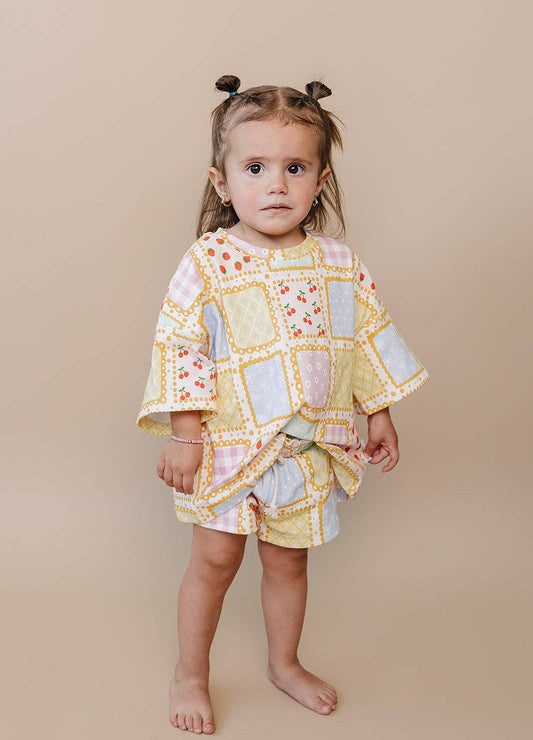 A toddler girl with her hair in two buns stands barefoot against a solid beige studio background, wearing a patchwork-print cotton outfit with a long-sleeve tunic top and matching shorts.