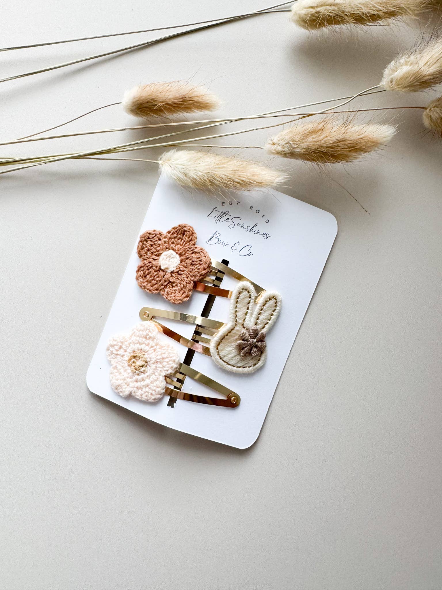 A 3-piece hair clip set on a white display card featuring a brown crochet flower, a cream crochet flower, and a cream embroidered bunny on gold-finish metal clips.