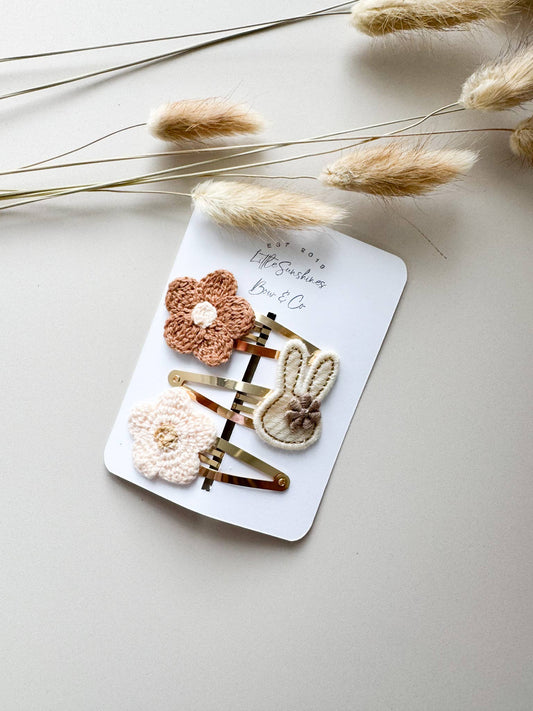 A 3-piece hair clip set on a white display card featuring a brown crochet flower, a cream crochet flower, and a cream embroidered bunny on gold-finish metal clips.