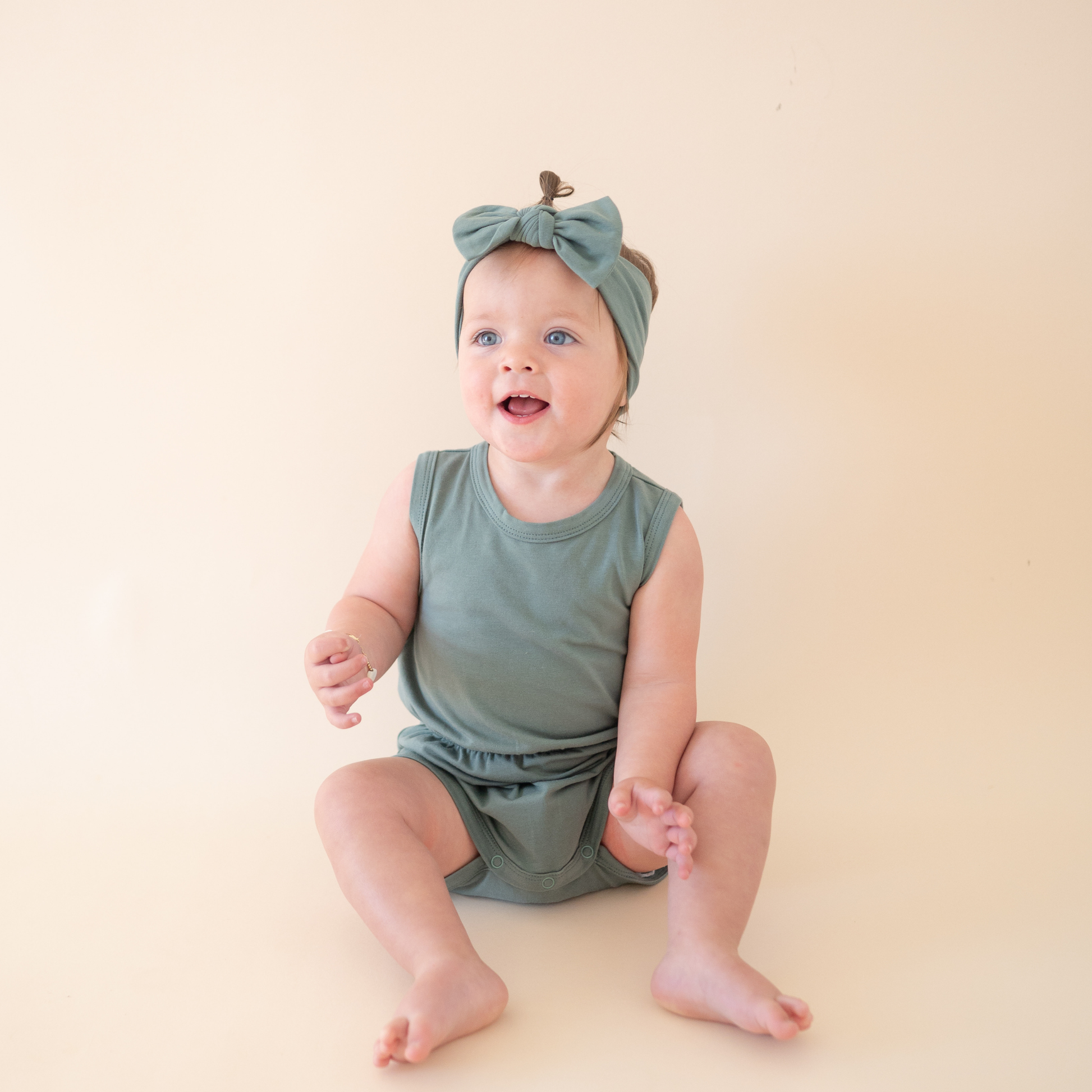 Baby sitting on a beige surface wearing a green outfit and headband.