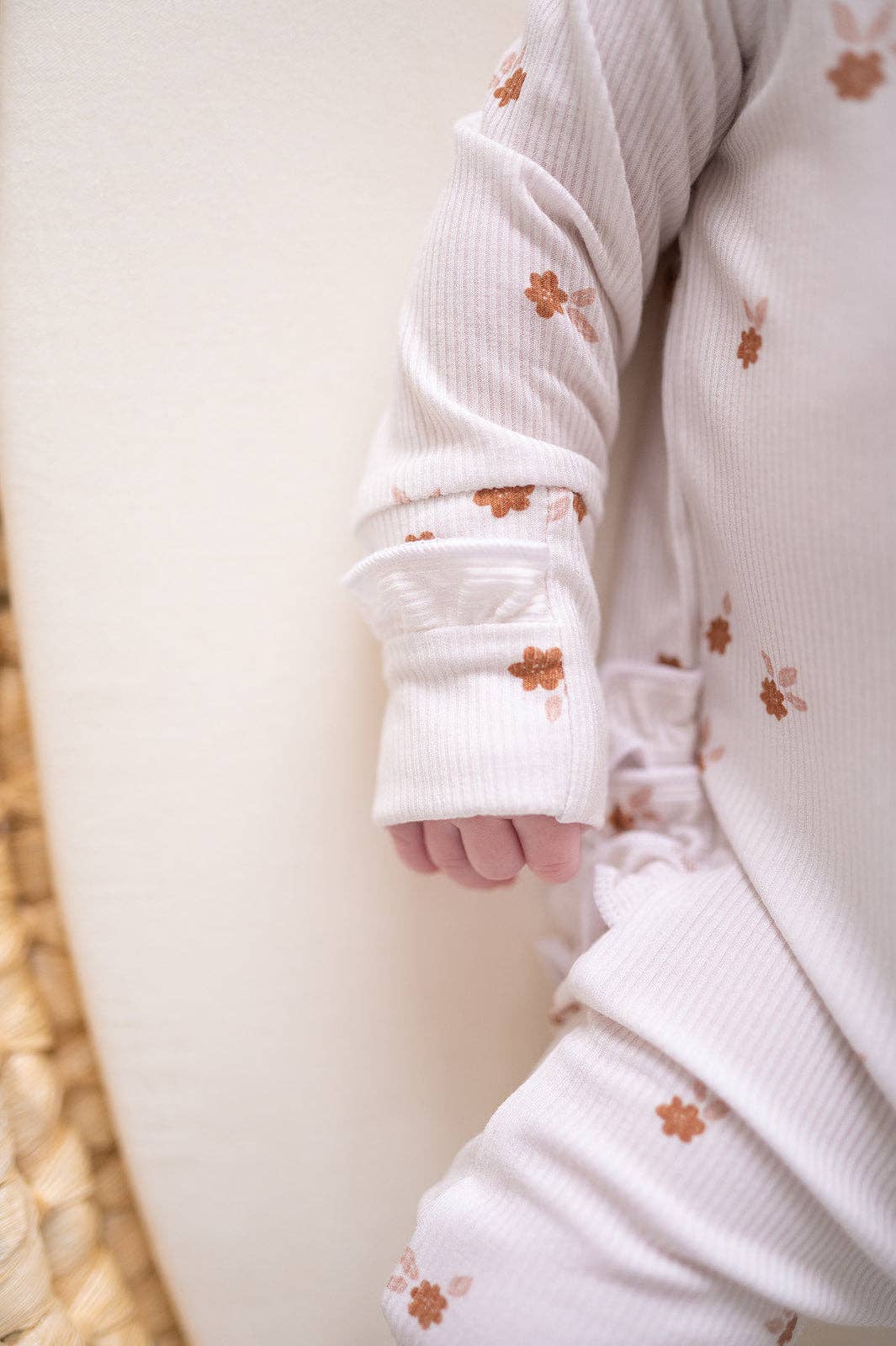 Close-up of a baby's hand and foot wearing a white outfit with orange floral patterns on a neutral background.