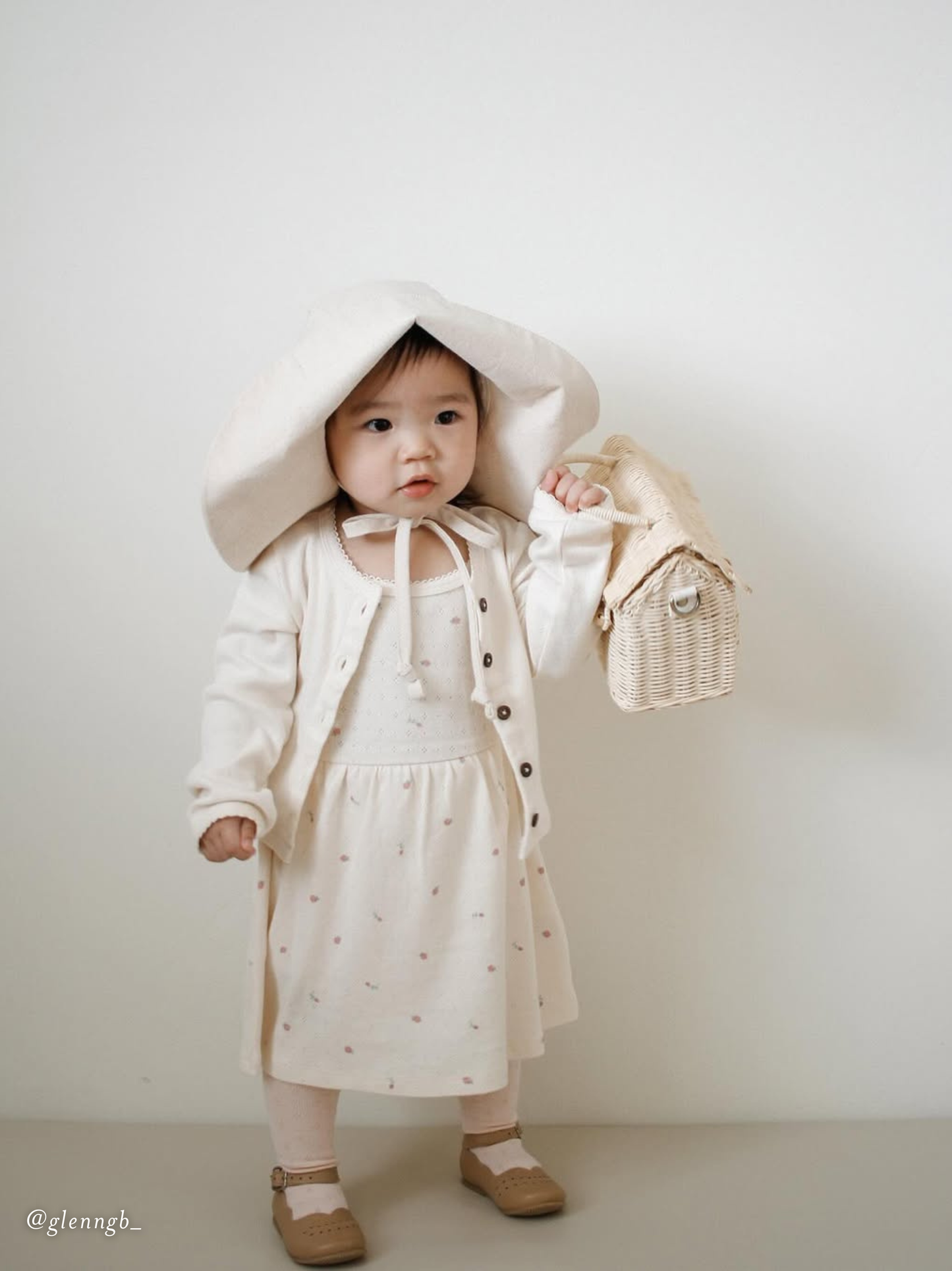 Child wearing a white hat with cardigan and holding a small bag against a plain background