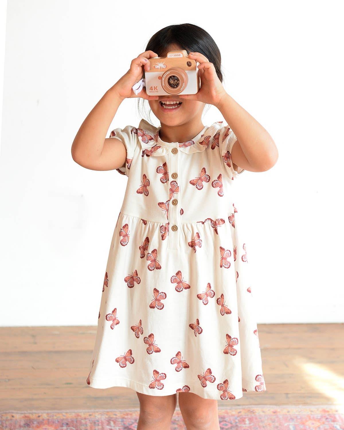 Child wearing a floral dress holding a toy camera against a white background