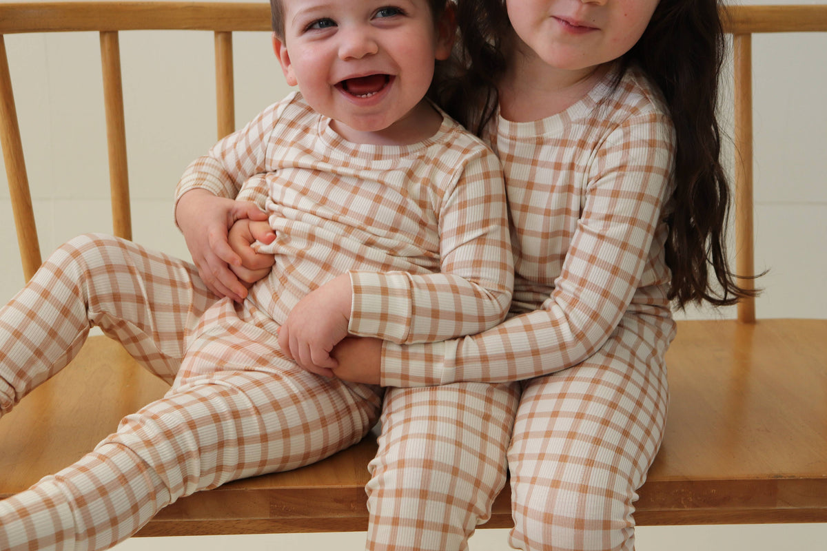 Two young children wearing matching beige and white gingham footed pajamas, sitting together on a wooden bench.