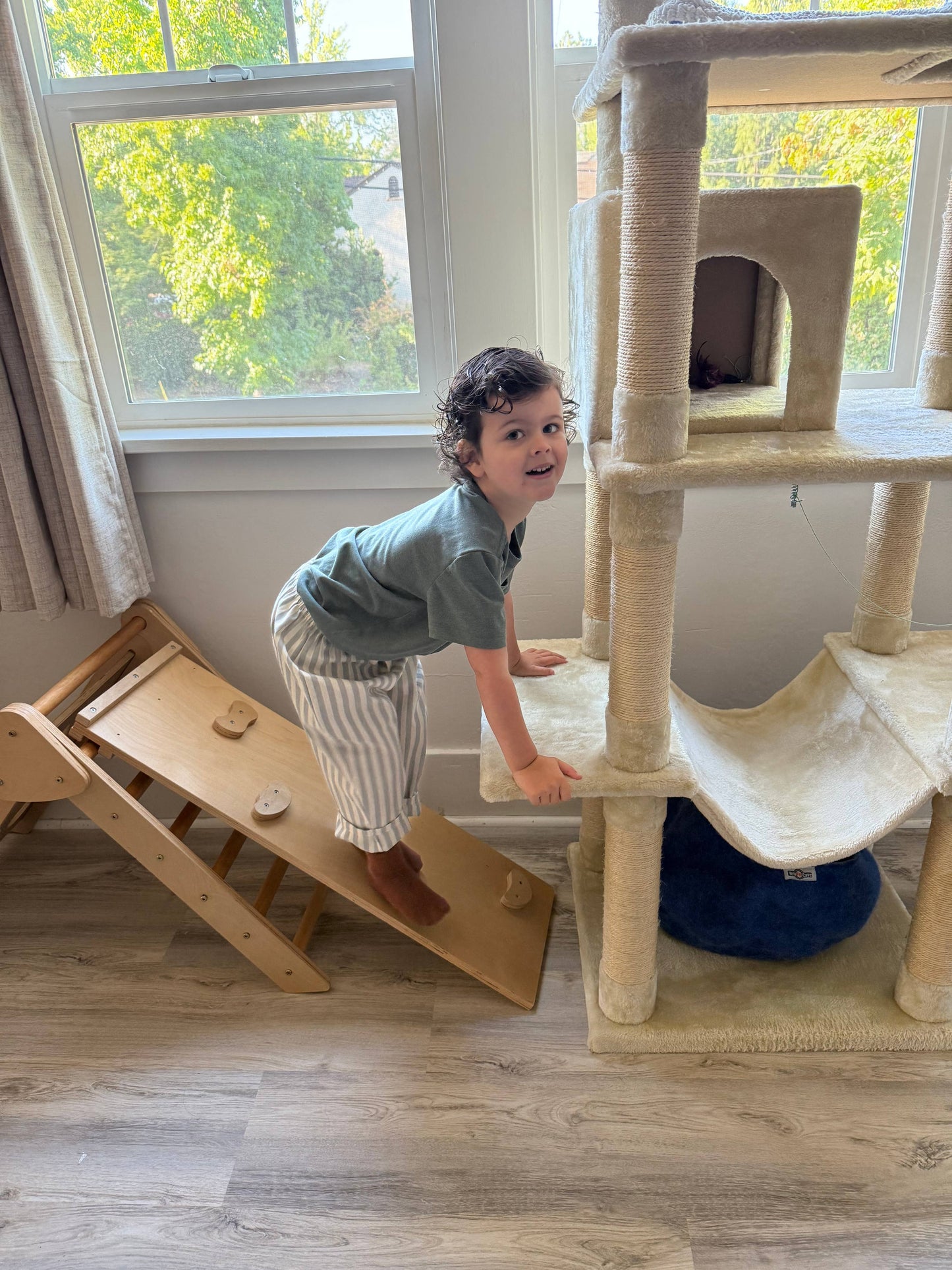 Child climbing a wooden climbing structure with a cat tree in a room with large windows.