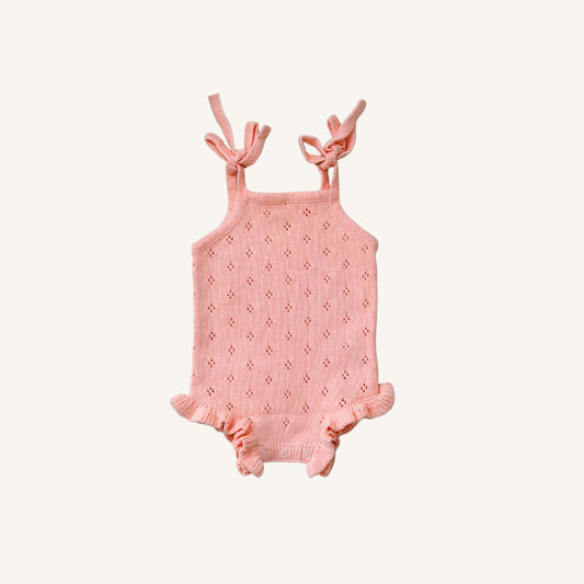 A light pink baby romper with a textured eyelet knit pattern, tie shoulder straps, and ruffled leg hems, displayed flat against a light cream background.