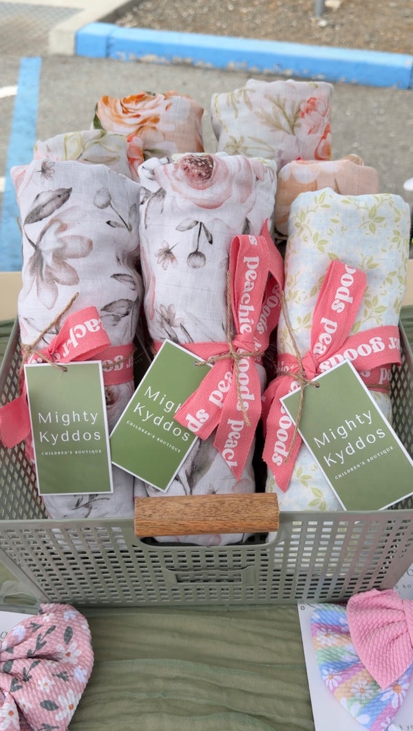 Rolled floral baby blankets in a grey basket with sage green Mighty Kyddos hang tags and pink ribbons