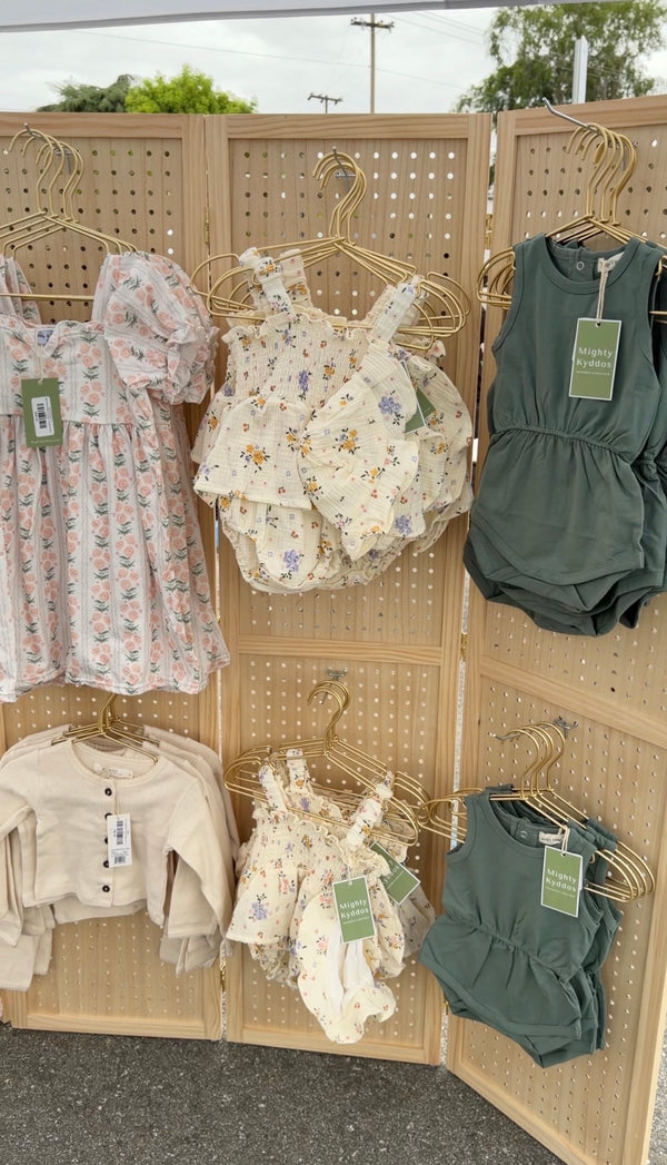 Children's boutique clothing display featuring floral dresses and sage green rompers on gold hangers against a wooden pegboard.