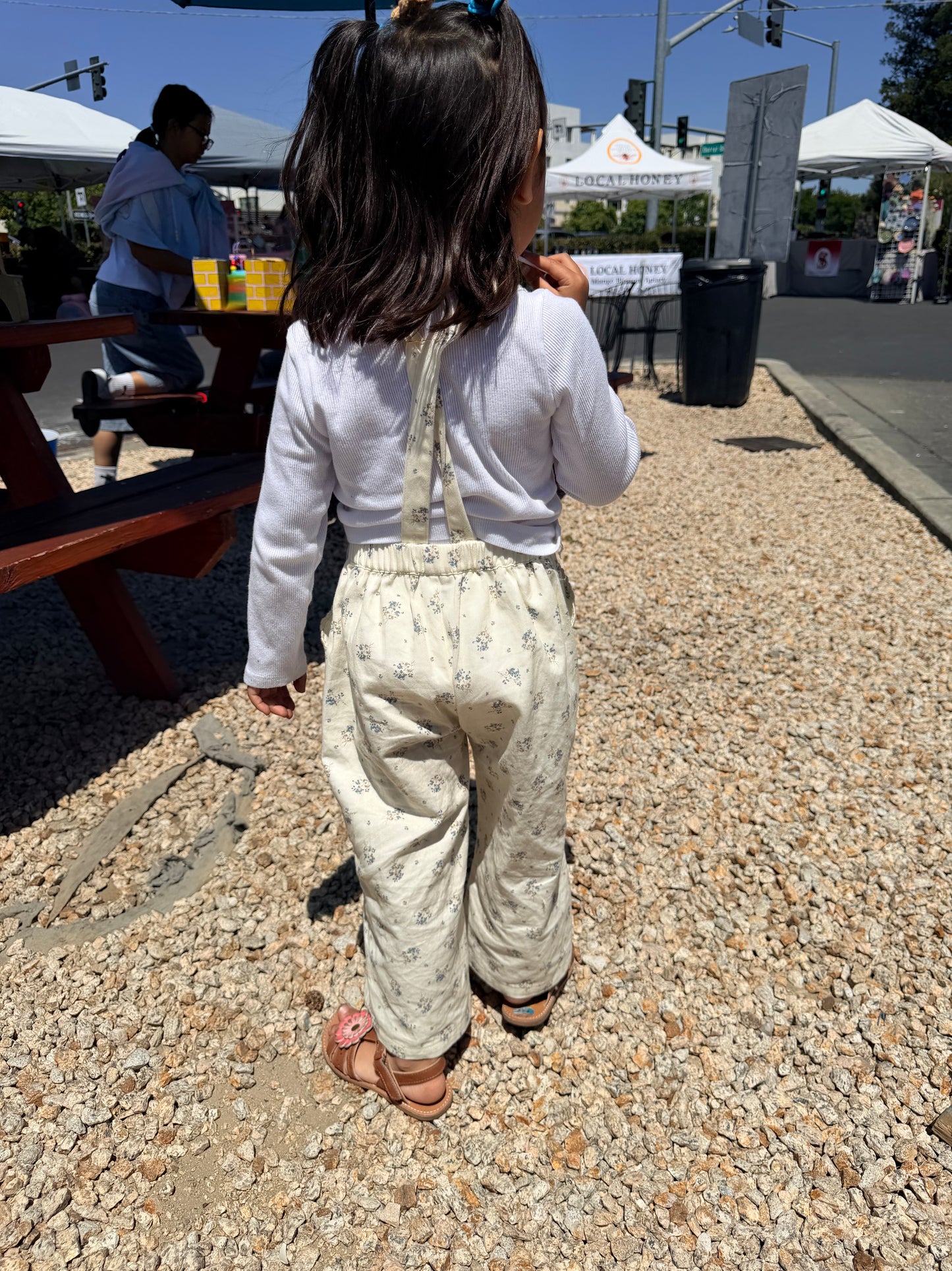 Child in a white shirt and patterned overalls walking on a gravel path with outdoor stalls in the background.