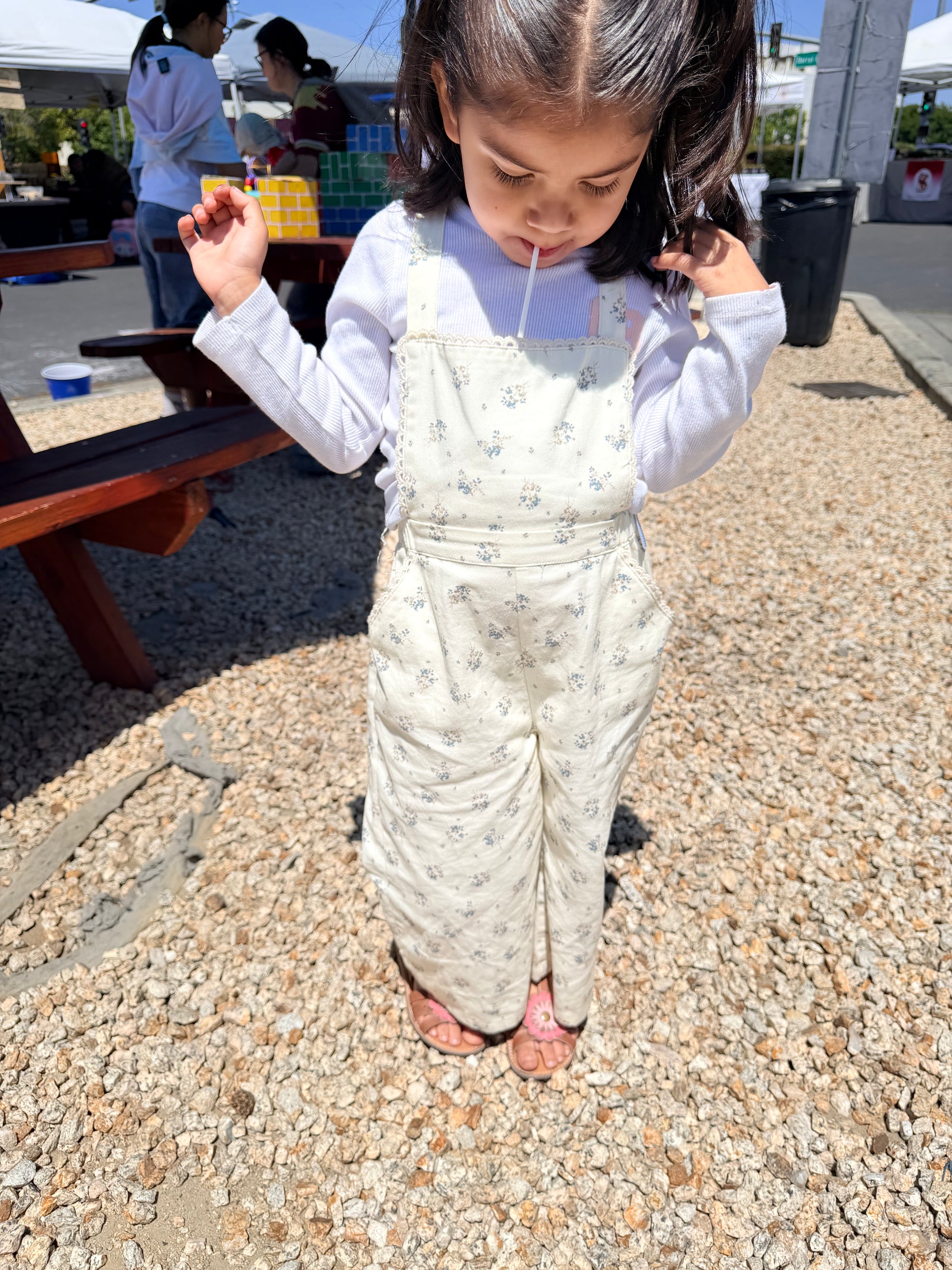 Child wearing a patterned overalls standing on a gravel surface with people and tents in the background.