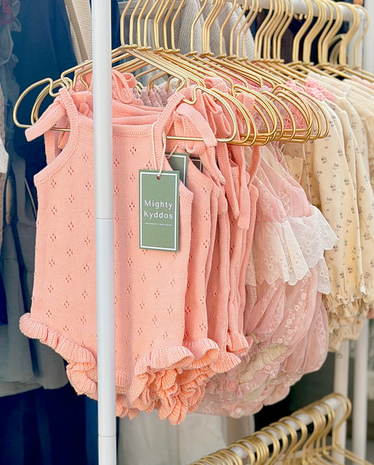 Pink pointelle knit baby rompers with shoulder ties and ruffled leg openings hanging on gold hangers on a white clothing rack, featuring a green Mighty Kyddos boutique tag.