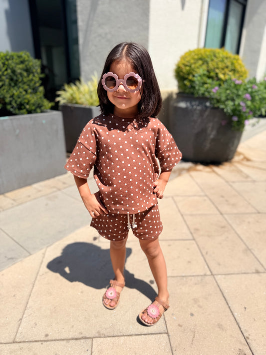 A young girl stands outdoors on a paved walkway wearing a matching brown and white polka-dot short-sleeve top and shorts set, pink sunglasses, and pink sandals.