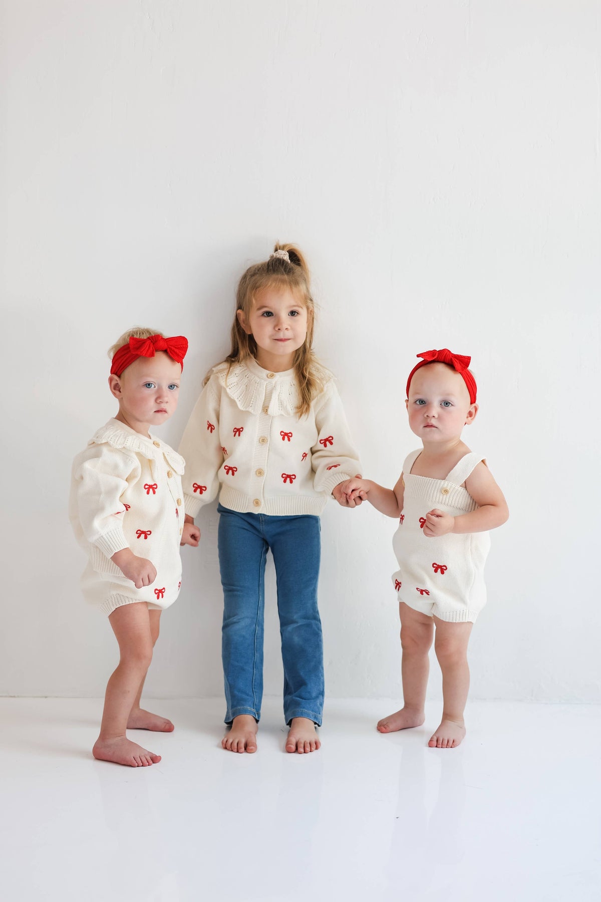 Three toddlers standing barefoot against a white studio wall, wearing matching cream romper outfits with small red bow details and red headbands.