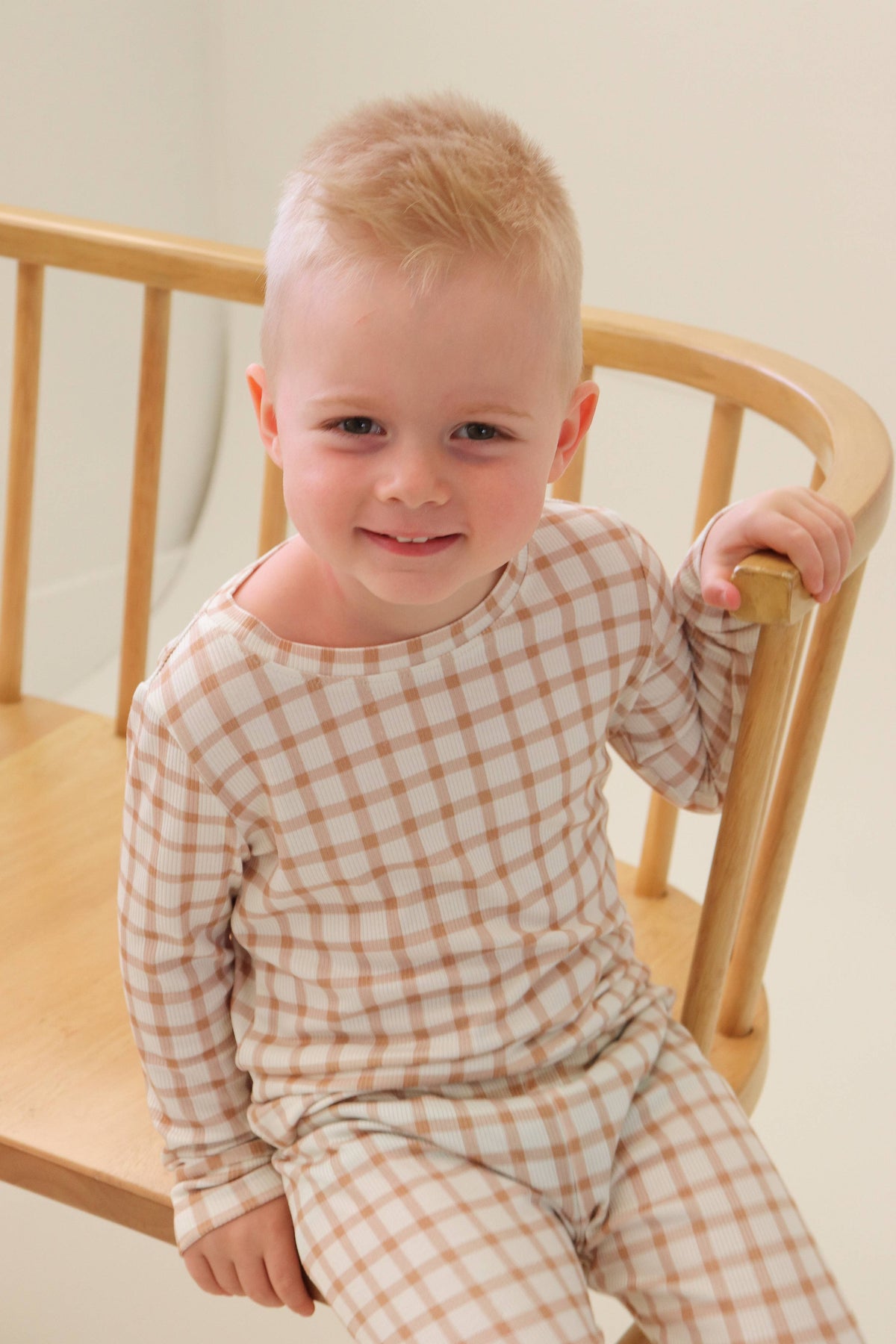 A smiling toddler with short blond hair sits on a light wood chair, wearing a beige and white gingham long-sleeve pajama set.