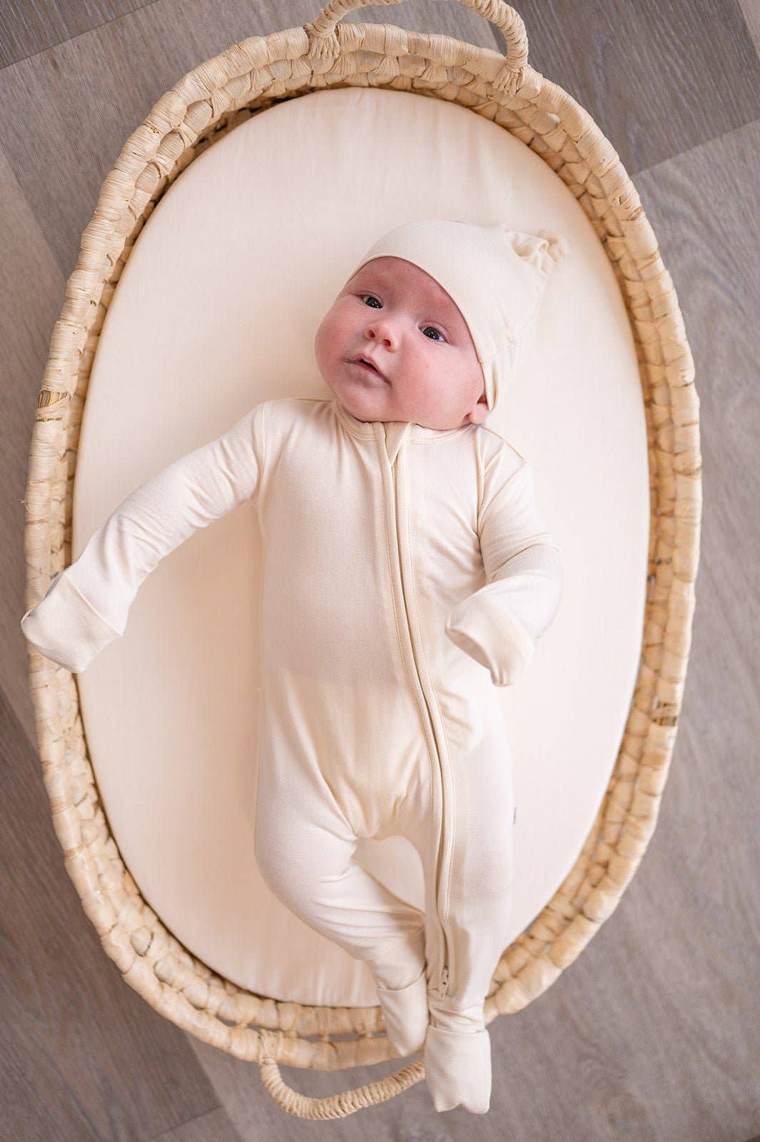 A peaceful lifestyle photograph of a baby lying in a woven wicker Moses basket. The baby is wearing a matching cream-white bamboo zipper romper and knotted beanie set. The scene is bright and minimalist, shot from a top-down perspective on a light-colored wood floor, highlighting the soft and premium feel of the neutral-toned fabric.
