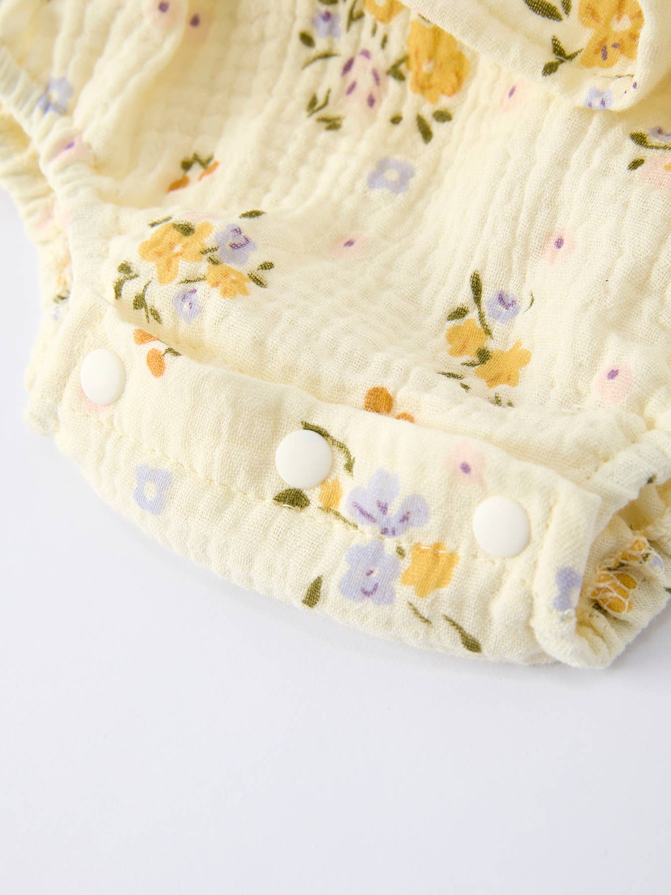 Bottom snap button closure of a cream floral baby romper made of textured cotton muslin on a white background