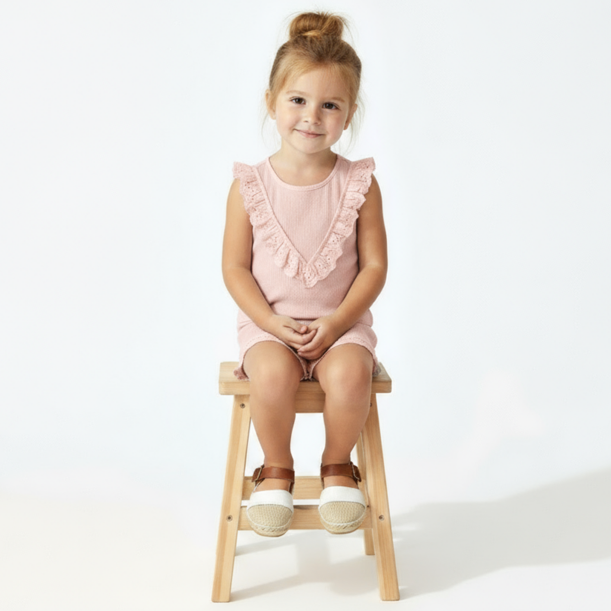 Girl in pink ruffled set sitting on a stool with a white background