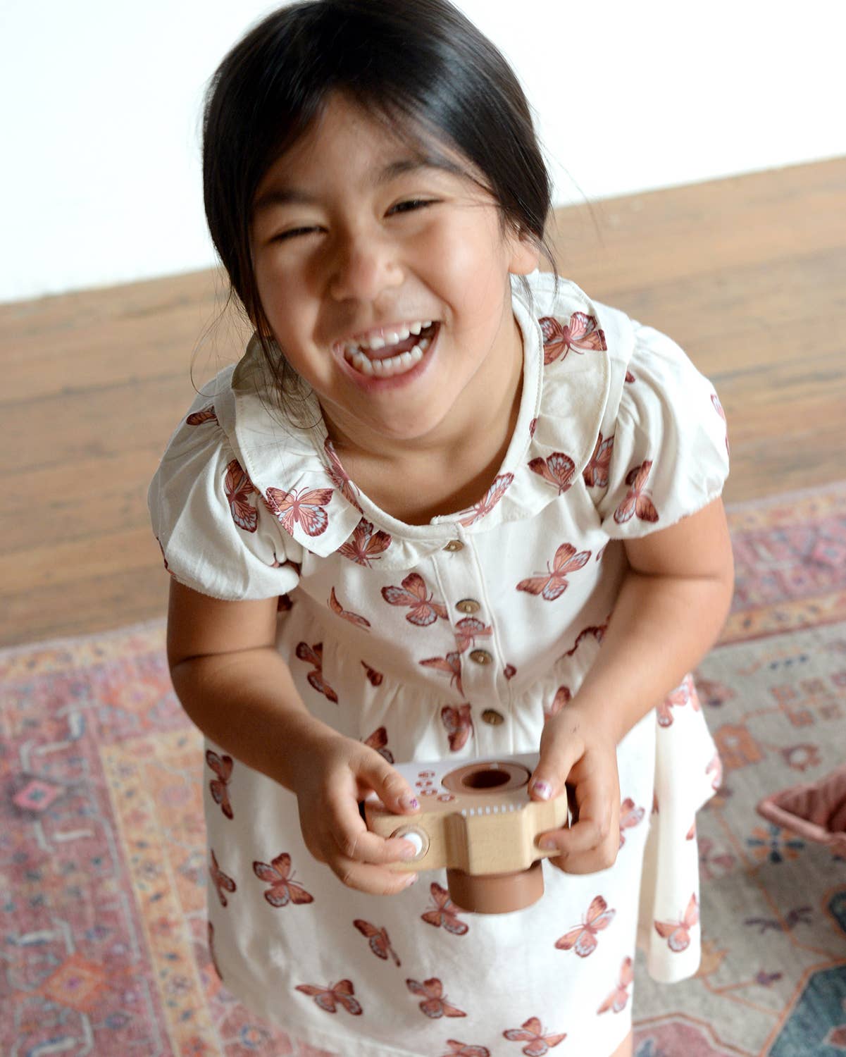 Child in a butterfly-patterned dress holding a wooden toy on a wooden floor.