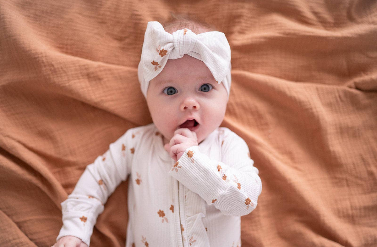 A baby wearing a white long-sleeve bodysuit and matching bow headband with small brown animal prints, lying on a textured rust-colored blanket.