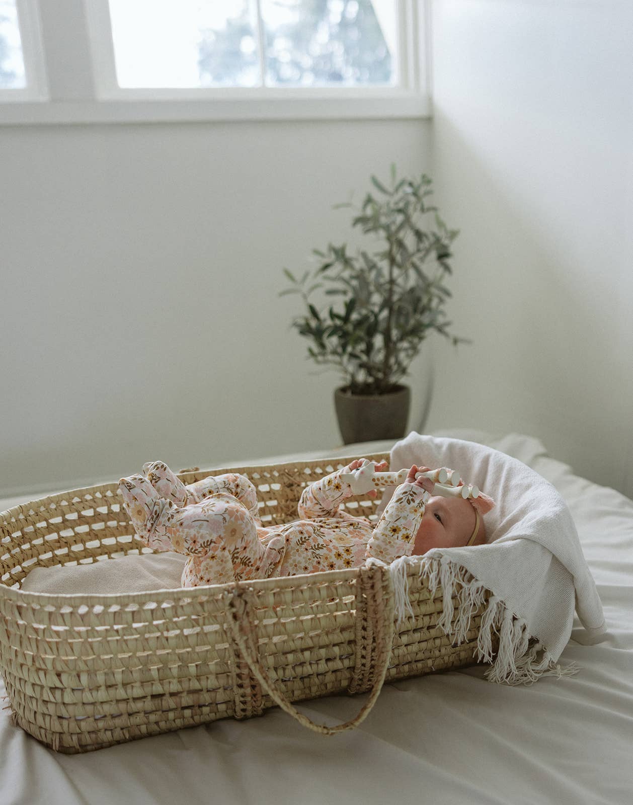 A serene lifestyle photograph of a baby lying in a woven wicker Moses basket. The baby is wearing a long-sleeved bamboo romper featuring a warm, vintage floral print in shades of peach and muted yellow. The basket is lined with a soft, fringed white textile and set against a clean, minimalist white wall. In the background, a small olive tree in a grey pot adds a touch of natural greenery. The entire scene is bathed in soft, bright natural light, emphasizing a peaceful and premium boutique feel.