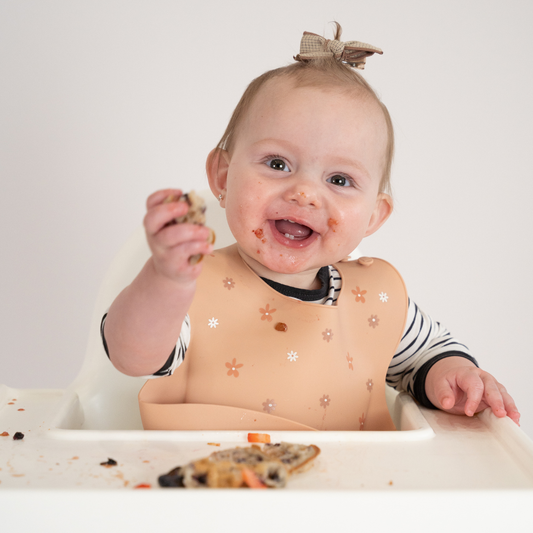 Baby in a high chair with food on face and bib, holding a cookie.