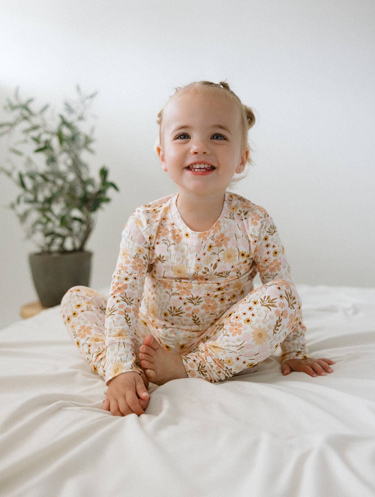 A bright, high-resolution lifestyle photograph of a smiling toddler sitting cross-legged on a clean, off-white textured sheet. The child is wearing a soft, long-sleeved bamboo two-piece pajama set featuring a vintage-inspired floral print with peach, muted orange, and mustard yellow wildflowers. In the soft-focus background, a small olive tree in a dark pot sits against a minimalist white wall. The scene is lit with soft, natural light, highlighting the gentle and premium quality of the fabric.