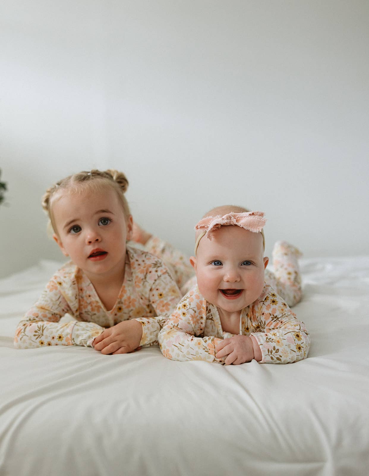 Two young children, a toddler and a baby, lie side-by-side on their stomachs on a clean white sheet. They are both wearing matching long-sleeved bamboo pajamas featuring a vintage-inspired floral print in shades of peach and muted yellow. The baby wears a soft pink bow headband and smiles at the camera, while the toddler looks off-camera with a curious expression.