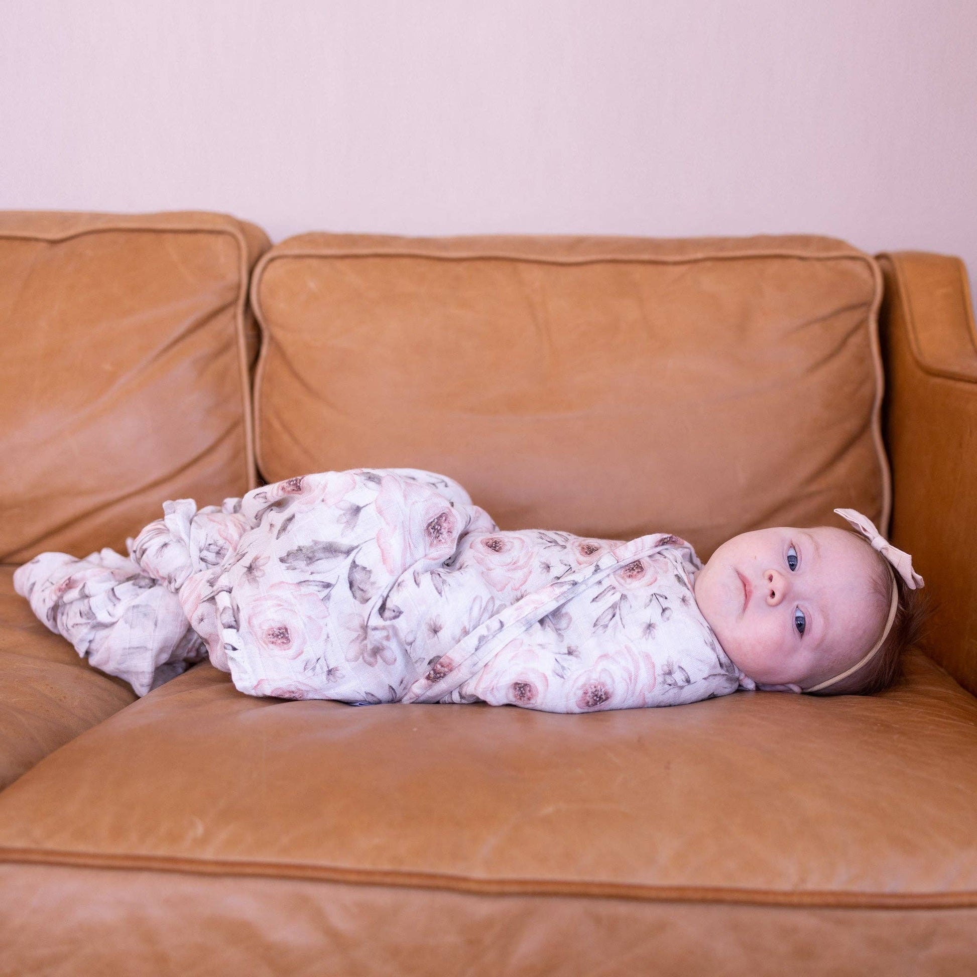 Baby swaddled in a floral blanket lying on a brown couch.