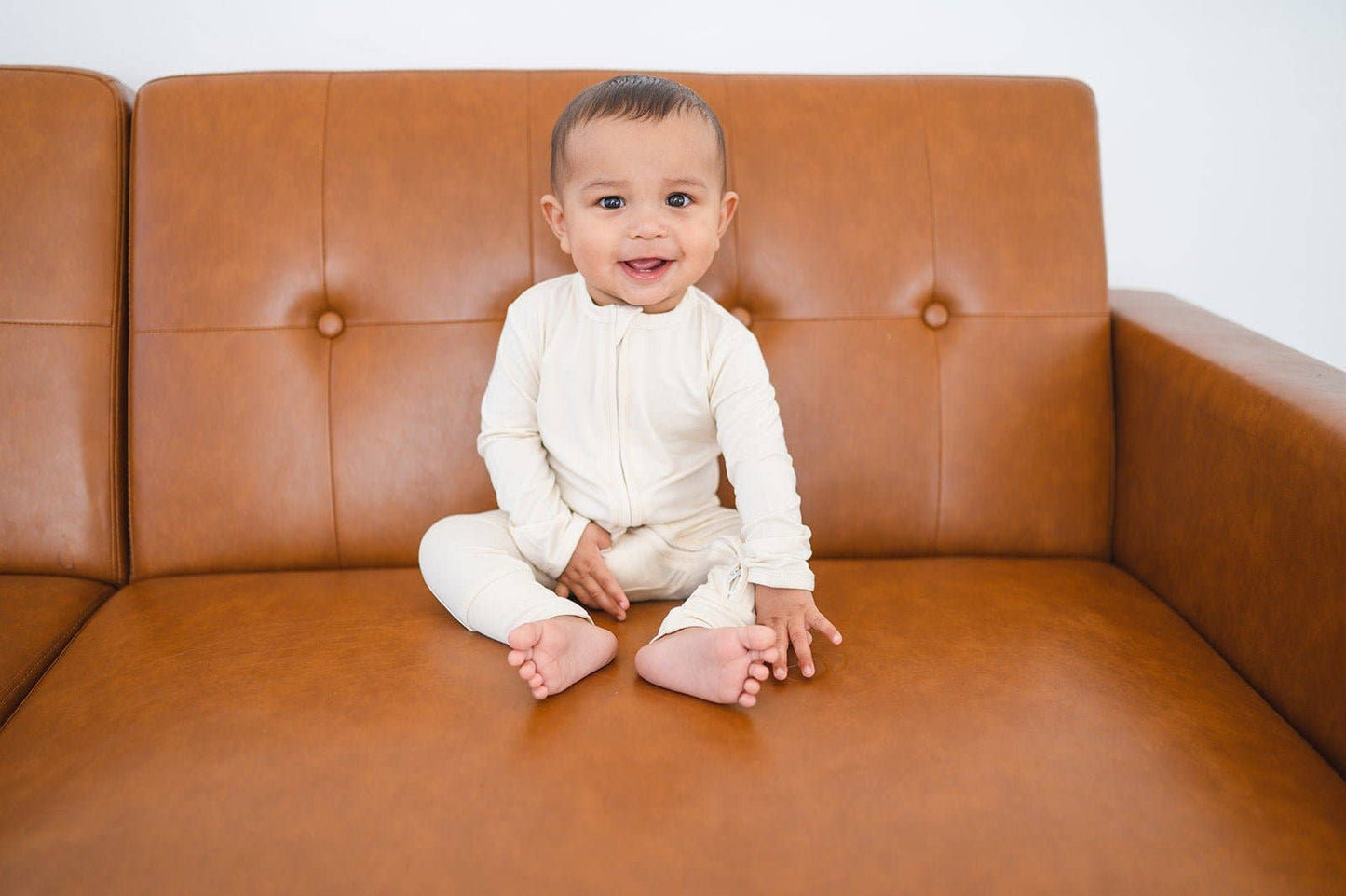 Baby sitting on a brown leather couch wearing a white onesie outfit.