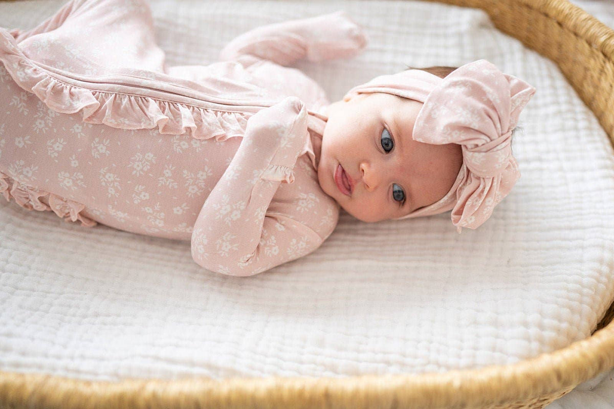 Baby in a pink outfit with bow headband lying in a crib.